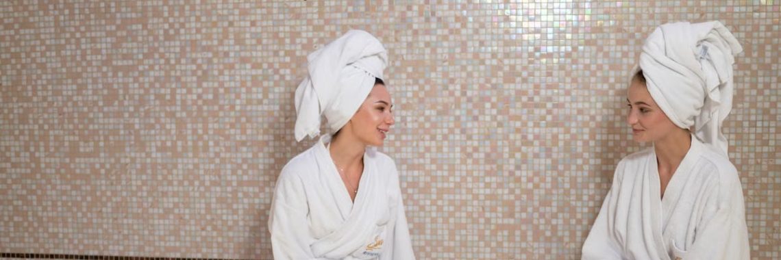 Two women in bathrobes and head towels enjoying a relaxing spa day with Turkish tea indoors.